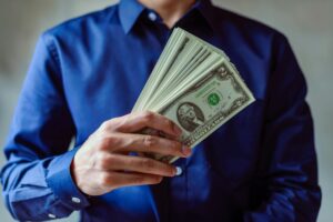 Close-up of a person in blue shirt holding multiple US dollar bills.