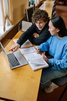Two students working together on a laptop at a desk in a library, smiling and engaging.